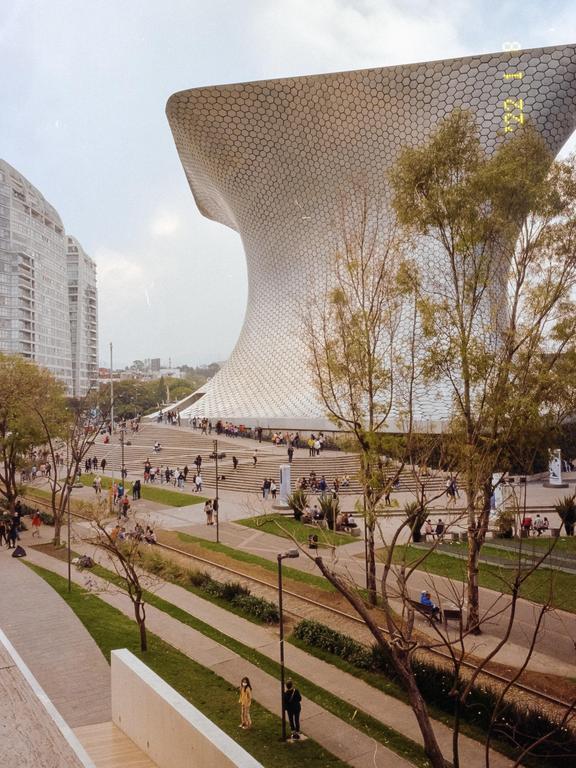 A large, modern, hexagonal building stands behind a park with trees, people, and train tracks.