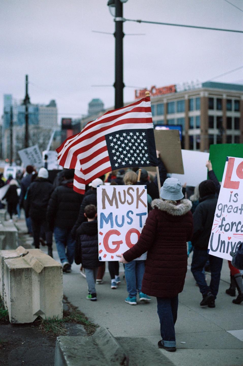 A crowd of people marches down a city street, some holding protest signs and an American flag.