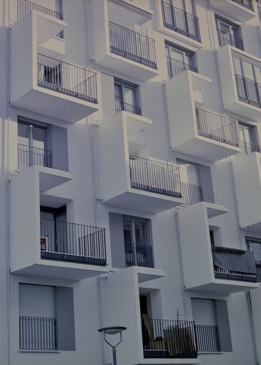 A modern building facade features numerous white balconies with black railings, some with windows and others with closed blinds.