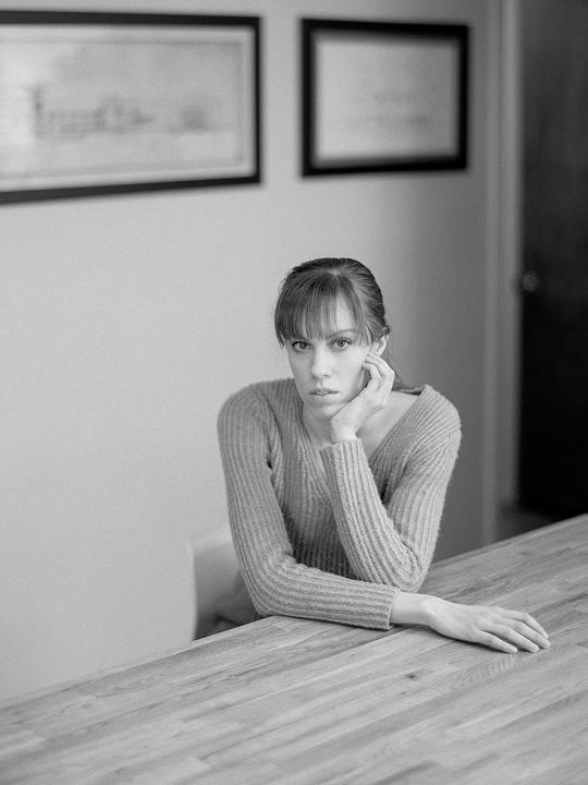 A woman with bangs and a ribbed sweater sits at a wooden table with her chin resting on her hand.