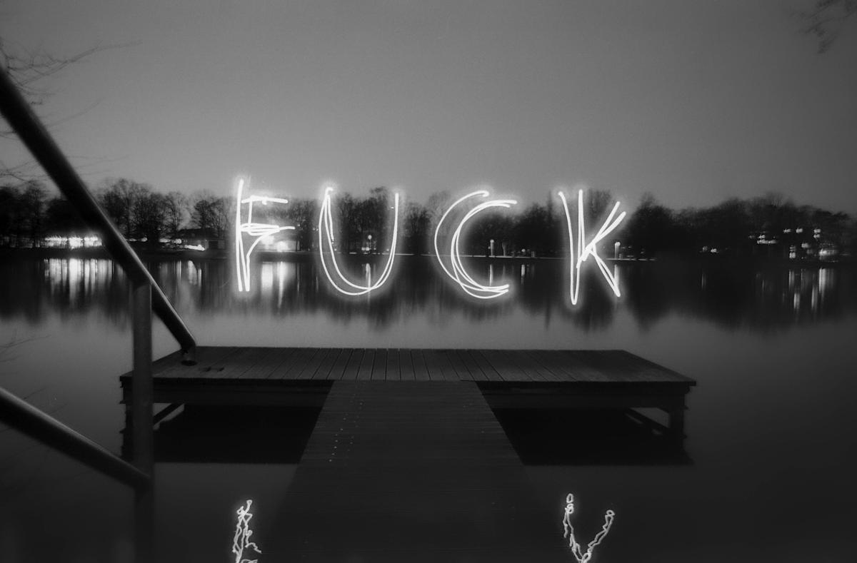 The word "FUCK" is spelled out in light over a body of water with a dock in the foreground.