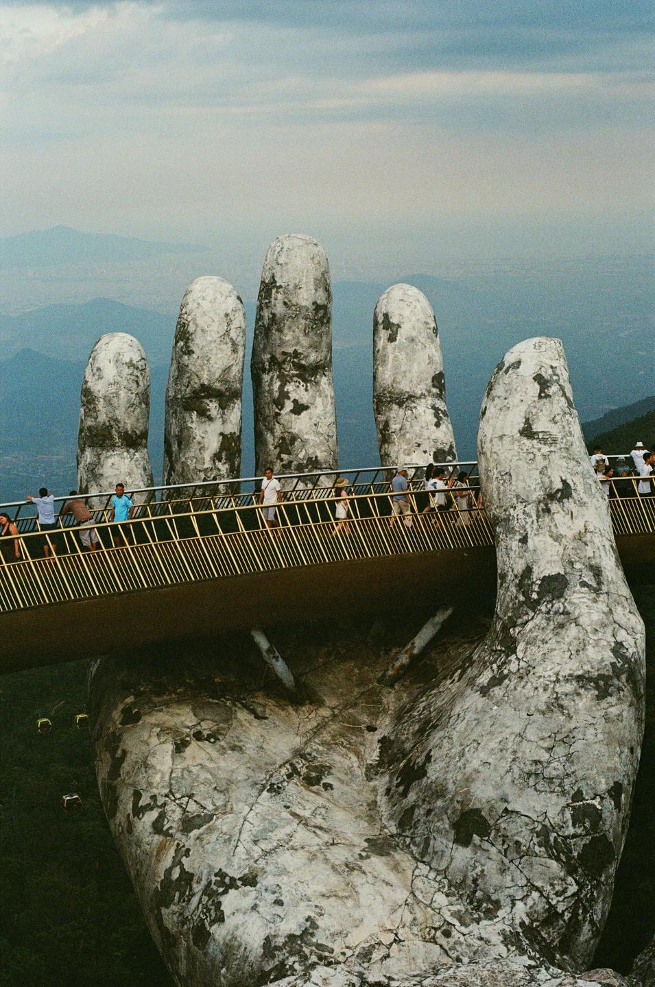 A large stone hand sculpture holds up a bridge with people walking on it, overlooking a hazy mountain landscape.