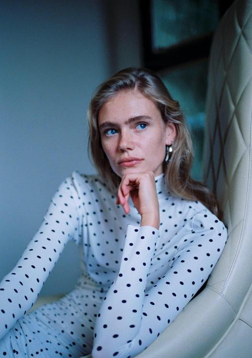 A woman with blonde hair and blue eyes wears a white polka dot top and rests her chin on her hand.
