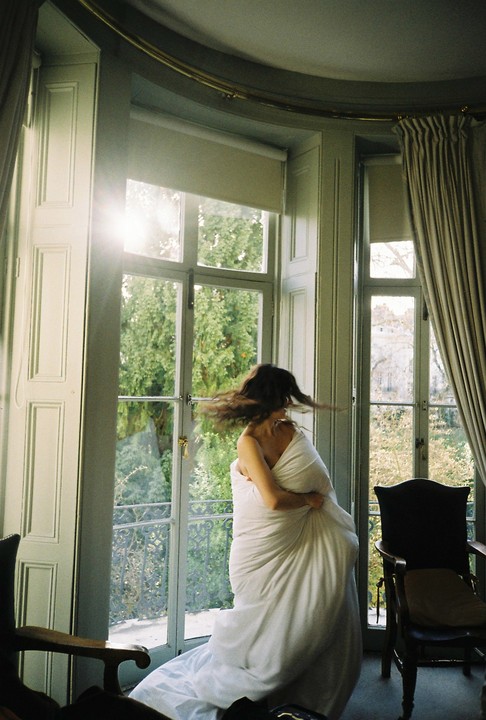 A woman wrapped in a white sheet spins in front of a large window with sunlight streaming through.