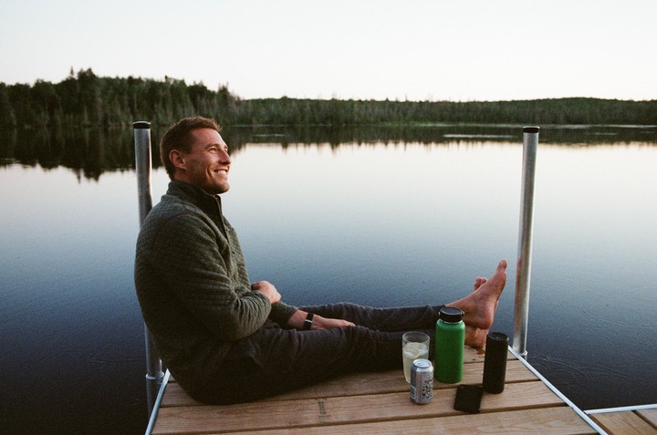 A man sits on a wooden dock with his feet stretched out, looking out at a calm lake surrounded by a forest.