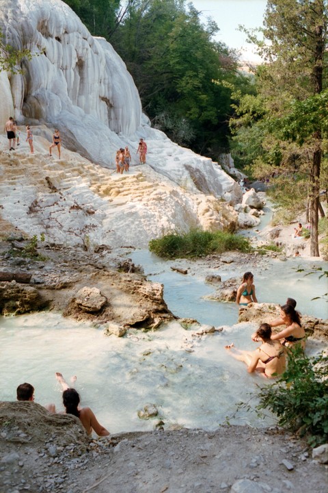 People are bathing in a light blue river with white rock formations and green trees on the riverbanks.