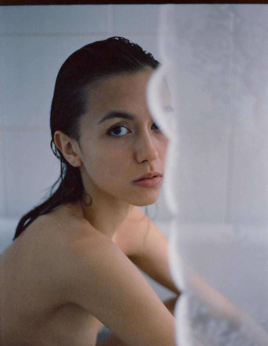 A woman with wet hair looks directly at the viewer from behind a shower curtain.