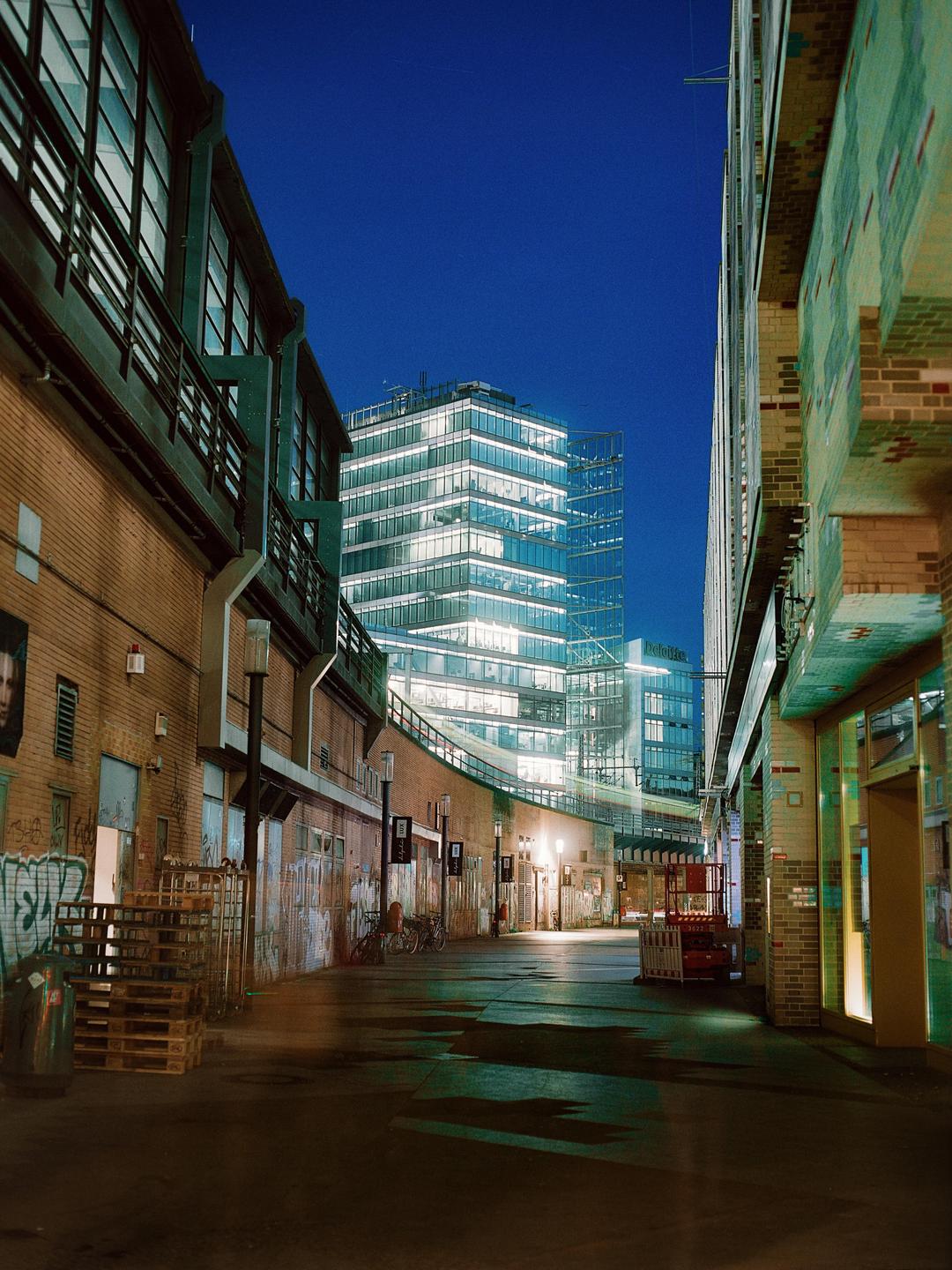 A narrow alleyway with brick buildings on either side leads to a brightly lit modern skyscraper under a dark blue night sky.