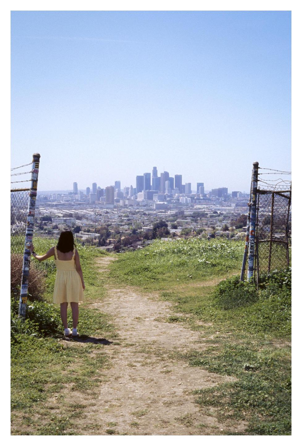 A woman in a yellow dress stands on a dirt path looking out at a city skyline.