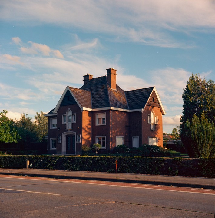 A large brick house with a dark roof and multiple chimneys stands behind a trimmed hedge, with a road in the foreground.