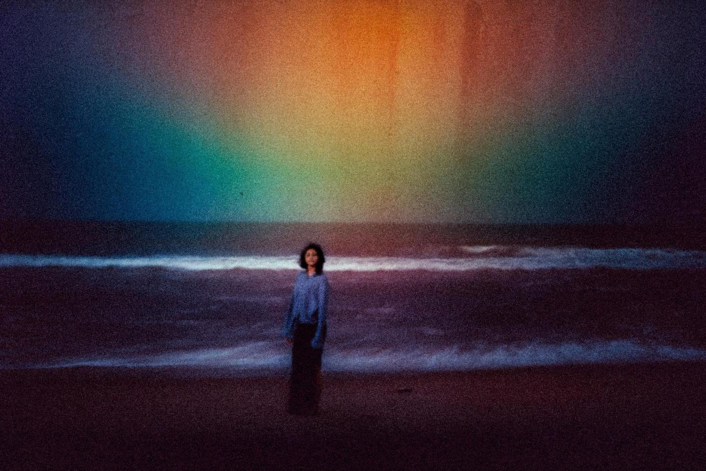 A woman stands on a beach with waves crashing behind her, under a sky with a rainbow-like light effect.