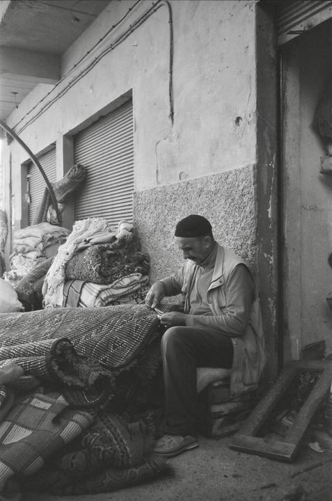 A man sits on a low stool, working on a rug in front of a building with closed shutters.