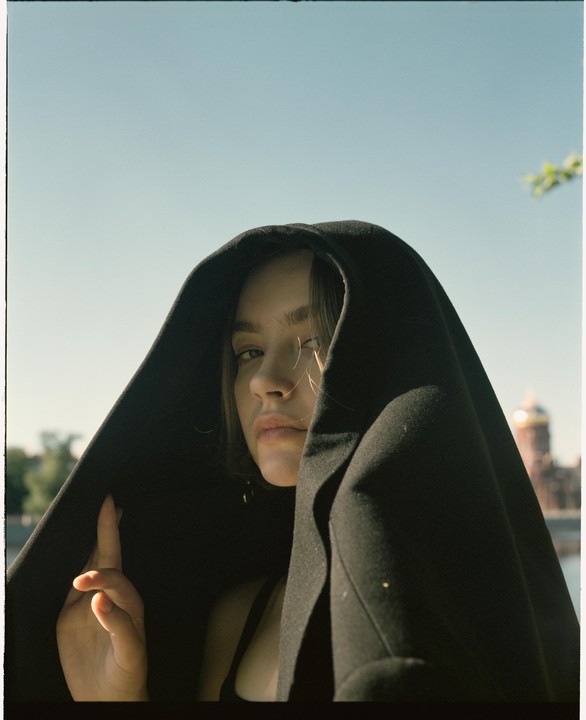 A woman with a black hood over her head looks at the viewer, with a golden dome building in the background.