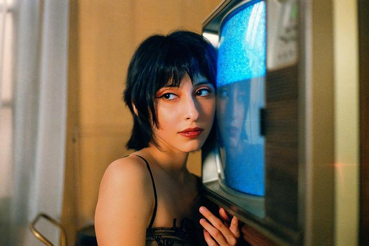 A woman with short dark hair and bangs looks to the side while leaning against a television with a blue screen.