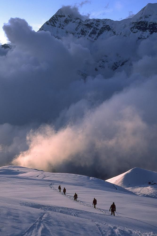 A group of people walk in a line through a snowy mountain landscape with clouds partially obscuring a large peak in the background.