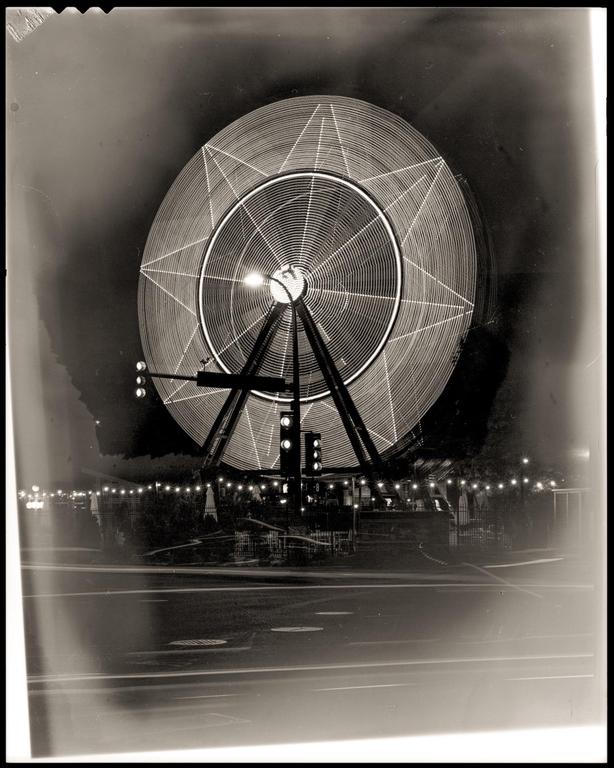 A long exposure captures the spinning lights of a ferris wheel at night, with light trails from passing vehicles on the street below.