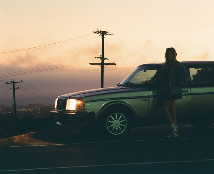 A woman stands next to a car with its headlights on, silhouetted against a hazy sunset sky with utility poles.