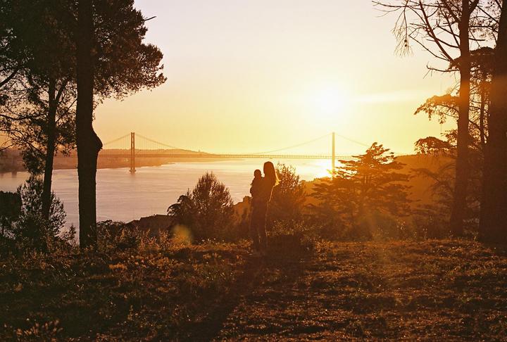 A woman holds a child on a hill overlooking a bridge over water at sunset.