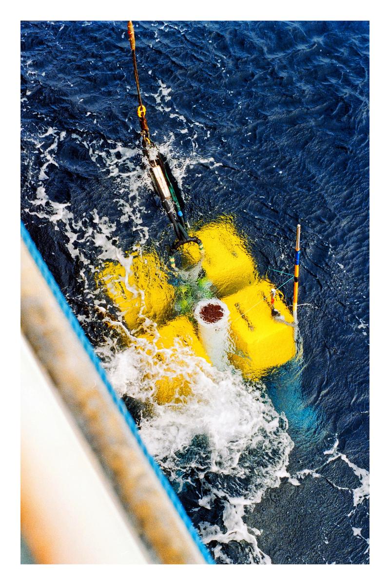 A yellow scientific instrument is being lowered into the dark blue ocean from a boat.