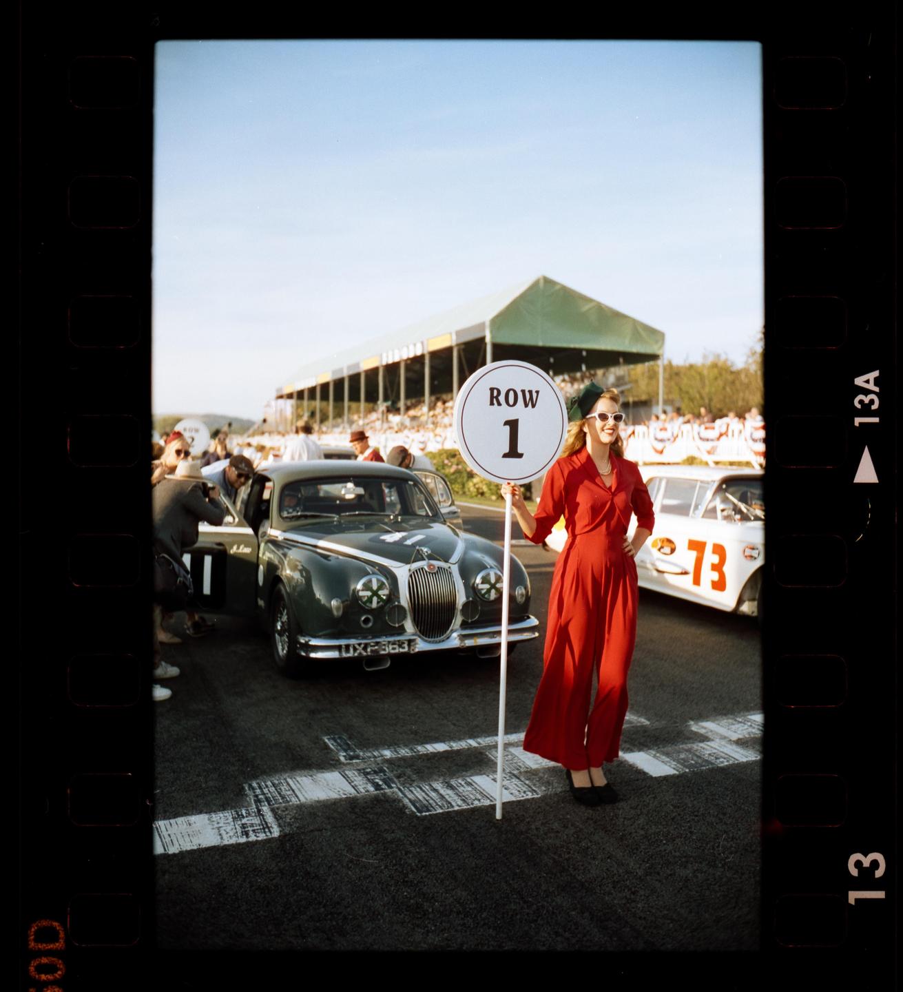 A woman in a red jumpsuit holds a "Row 1" sign on a racetrack next to a vintage car.