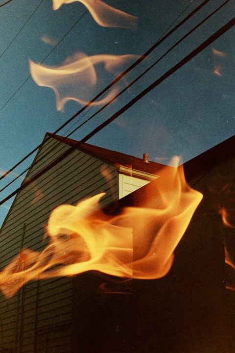 Flames are superimposed over a house with power lines against a blue sky.