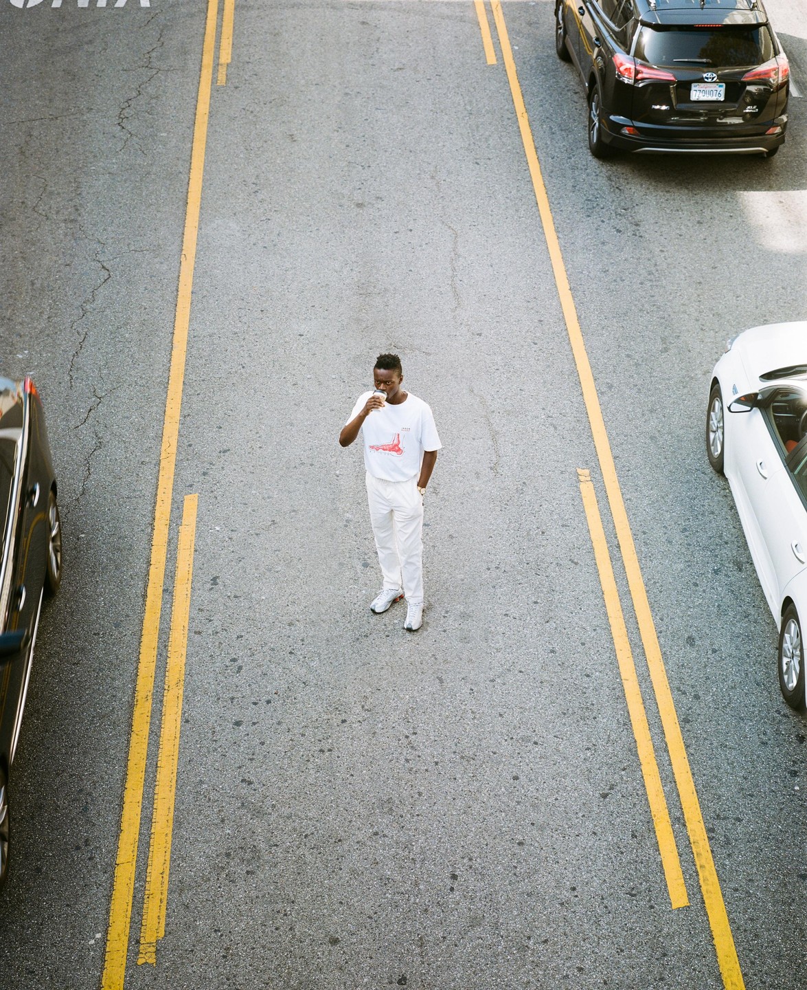 A man in white clothing stands in the middle of a street with cars on either side.