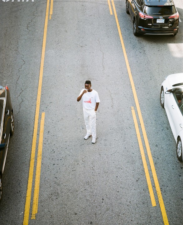 A man in white clothing stands in the middle of a street with cars on either side.