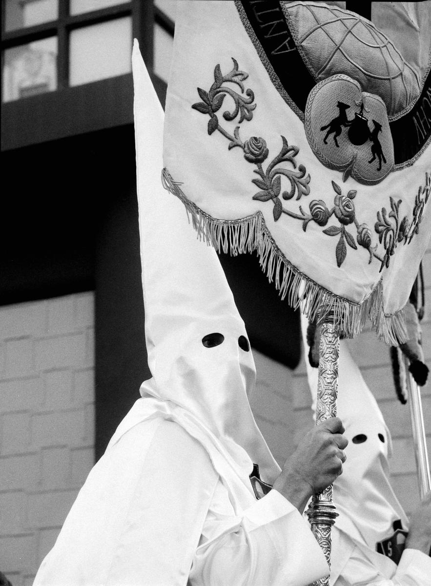 A person in a white hooded robe holds an ornate staff with a fringed banner.