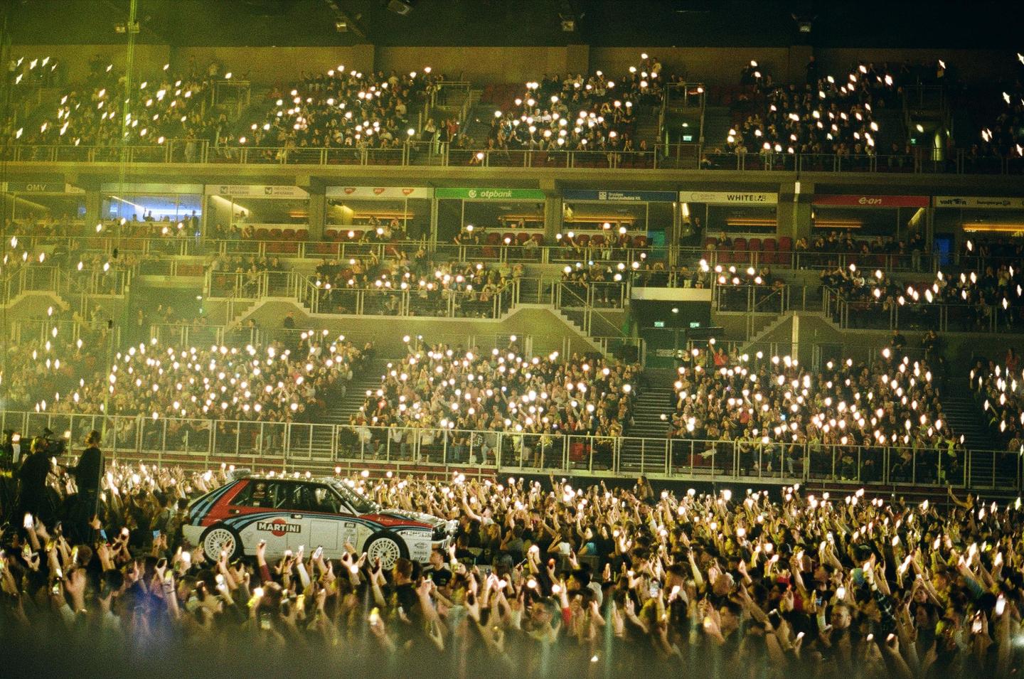 A rally car is surrounded by a crowd of people holding up lights in a stadium at night.
