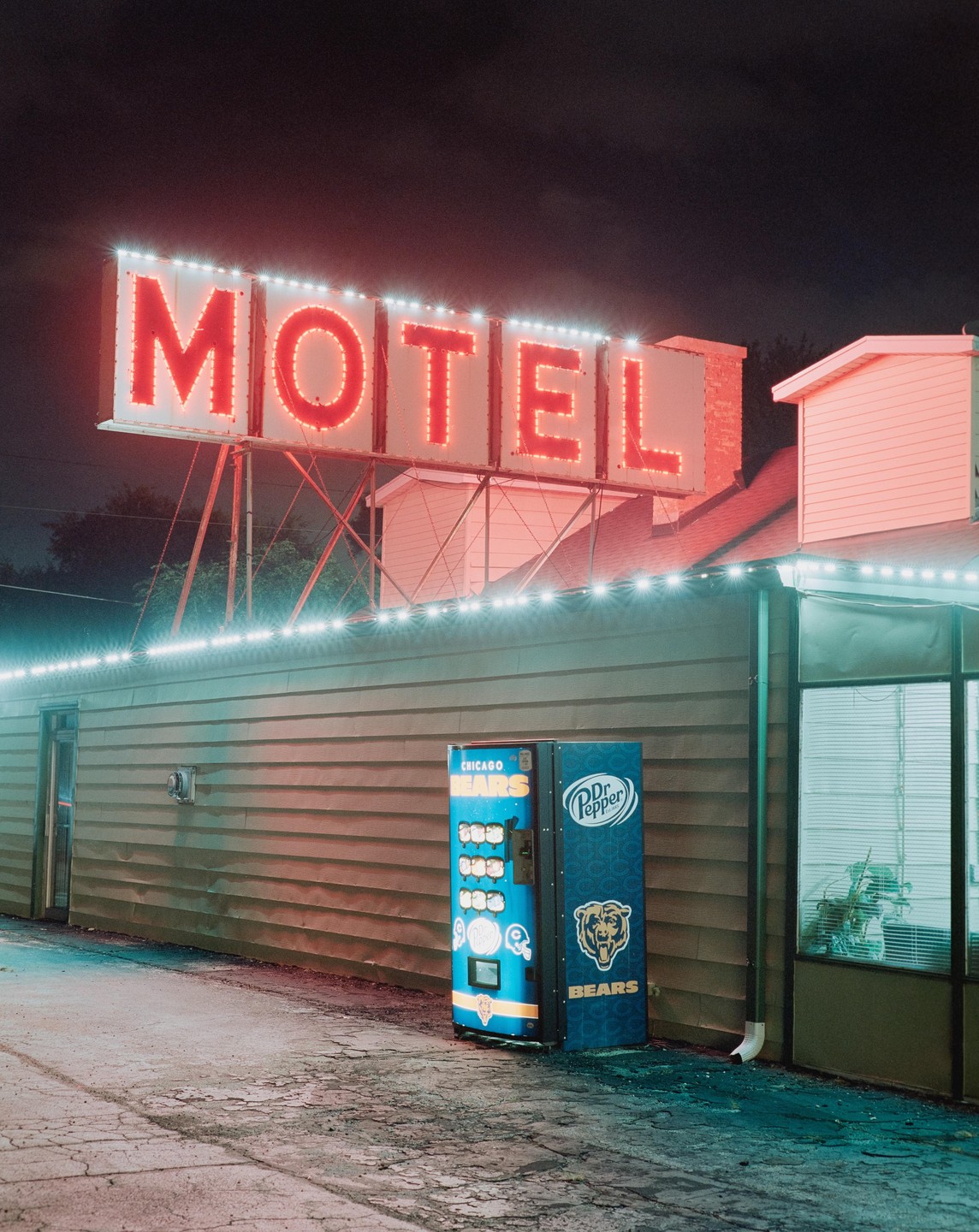 A neon motel sign glows red above a blue vending machine at night.