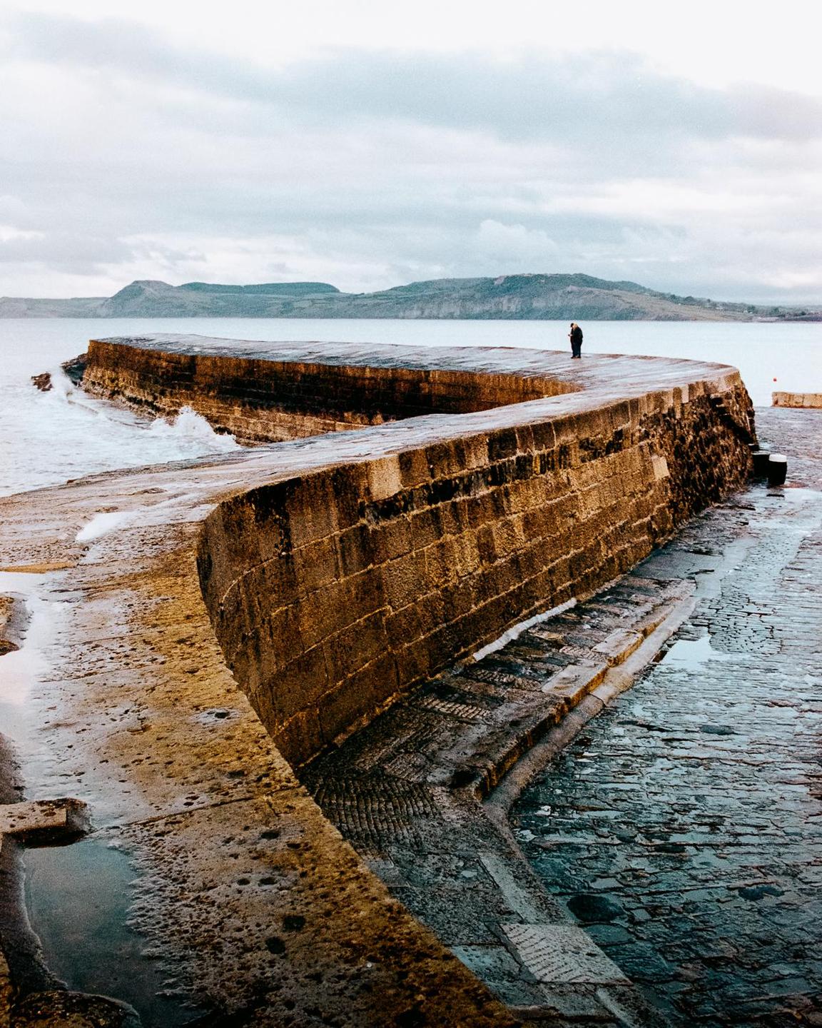A person stands on a curved stone pier with mountains in the background.