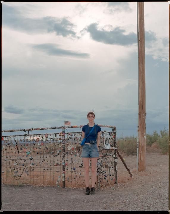 A woman stands in front of a fence covered in padlocks under a cloudy sky.