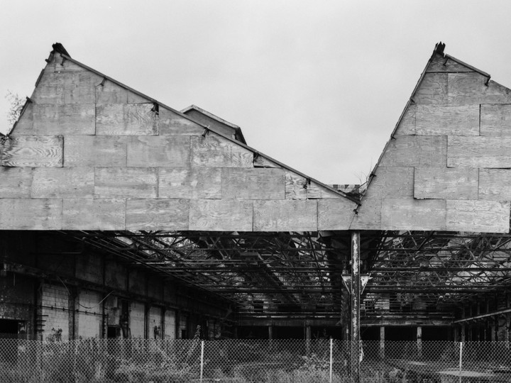 A chain link fence stands in front of an abandoned industrial building with a partially collapsed roof.