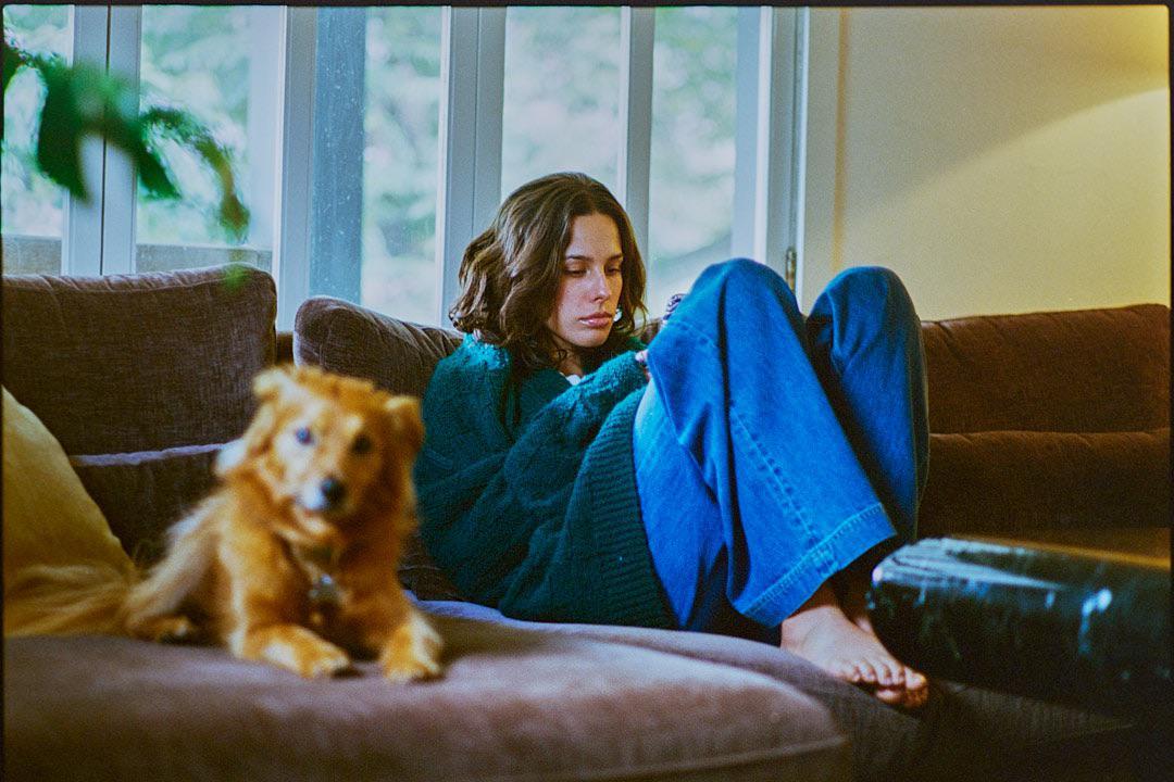 A woman in a green sweater and blue jeans sits on a brown sofa next to a small dog.