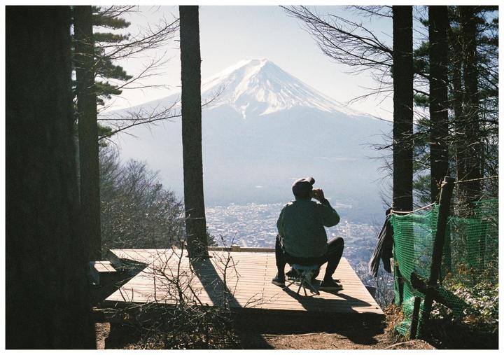 A man sits on a wooden platform looking out at a snow-capped mountain and a city below, framed by trees.