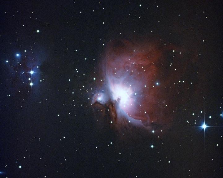 A bright nebula with red and blue hues is surrounded by numerous stars in a dark sky.