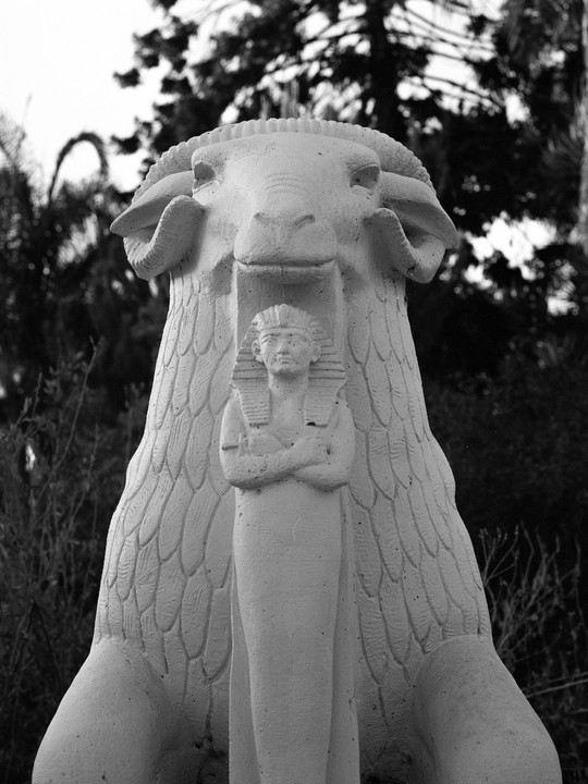 A statue of a ram with a pharaoh carved into its chest stands in front of trees.