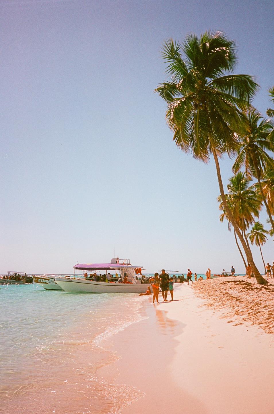 A boat is docked on a sandy beach with palm trees and people walking along the shore.