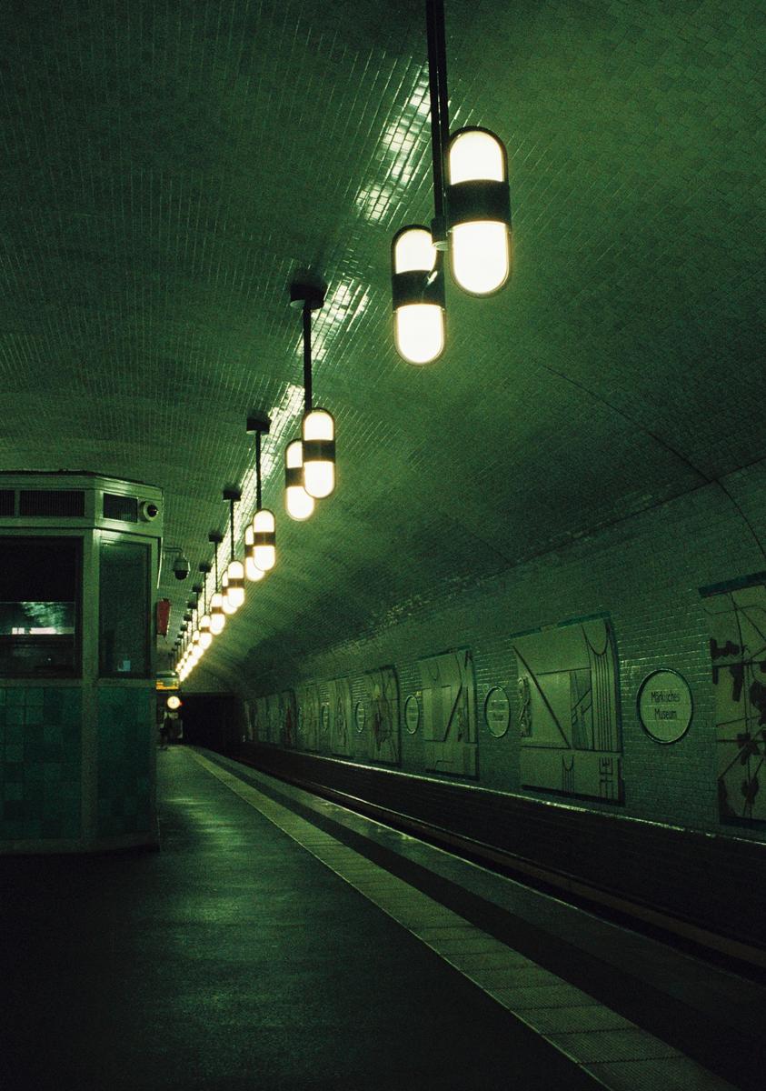 A subway platform with a tiled, arched ceiling is illuminated by a row of capsule-shaped lights.