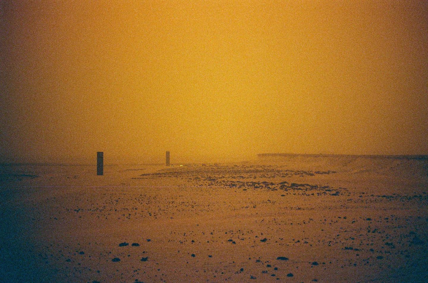 Two dark rectangular posts stand in a vast, hazy, orange-toned landscape with a distant horizon.