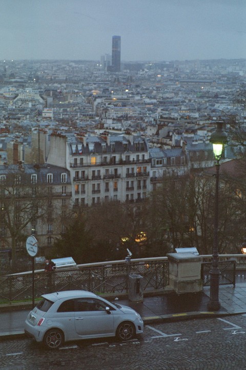 A small car is parked on a cobblestone street overlooking a city with many buildings and a tall skyscraper in the distance.