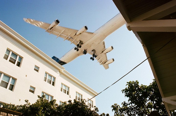 An airplane flies low over a white building and trees against a clear blue sky.