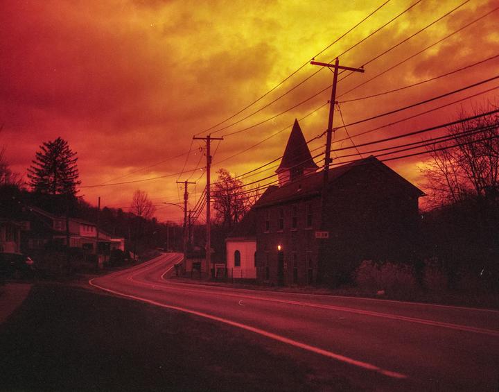 A winding road leads past a dark building with a steeple under a dramatic red and yellow sky.