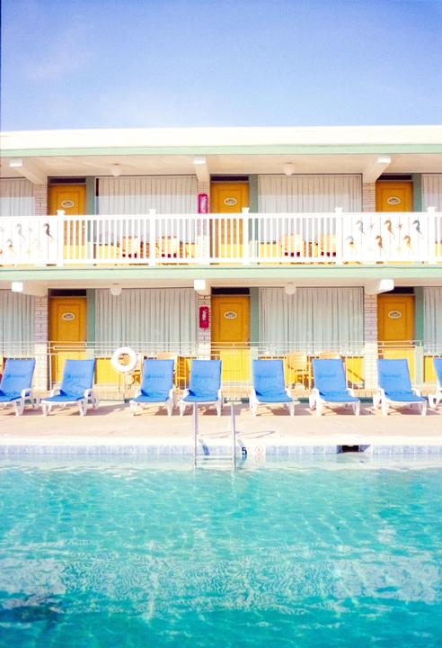 A swimming pool with blue lounge chairs in front of a two-story motel with yellow doors.