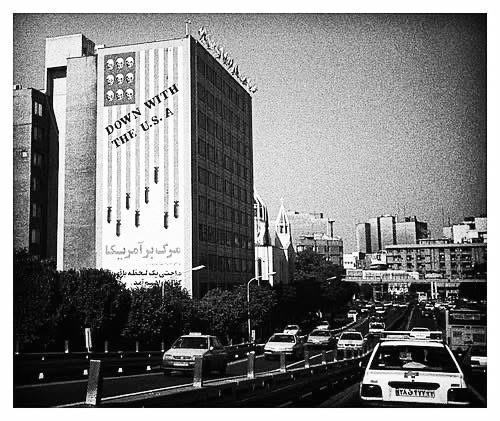A large mural on the side of a building depicts an American flag with skulls and bombs, overlooking a busy street with cars.