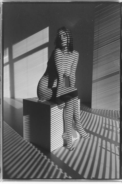 A woman sits on a cube with shadow stripes from window blinds covering her body and the room.