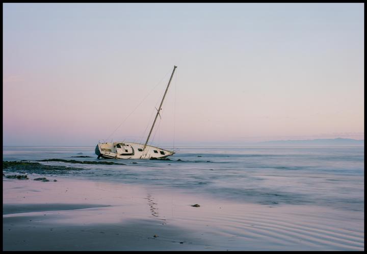 A wrecked sailboat sits on a rocky beach with mountains in the distance under a pastel sky.
