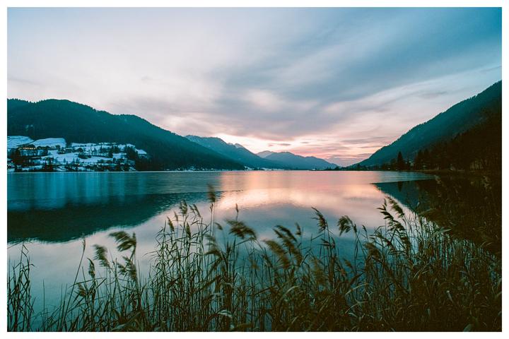 Reeds grow in the foreground of a lake reflecting a colorful sunset and surrounded by snow-covered mountains.