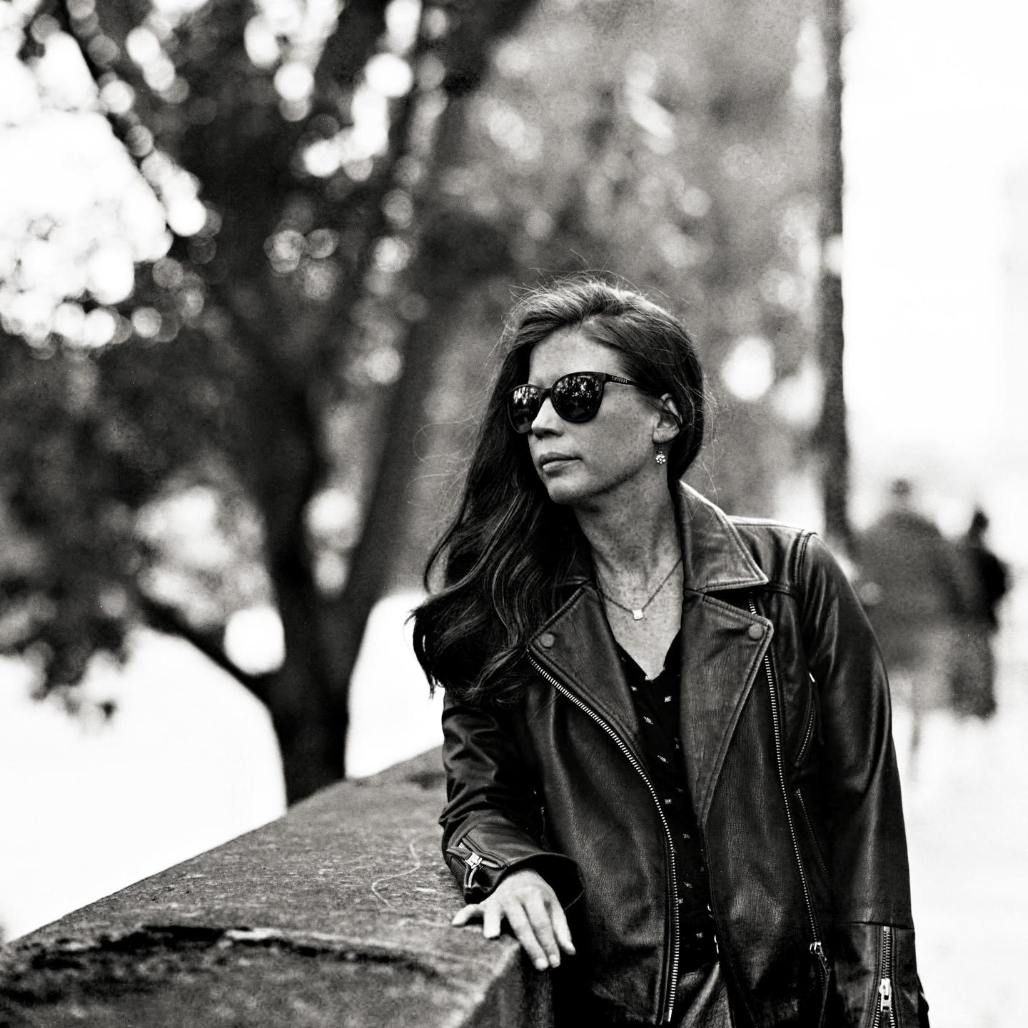 A woman wearing sunglasses and a leather jacket leans on a stone wall with trees in the background.