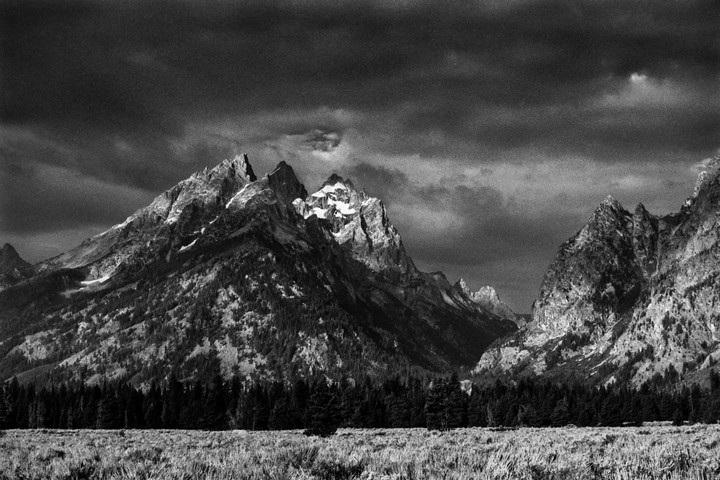 A dramatic black and white landscape shows mountains with snow-capped peaks under a cloudy sky, with a forest and field in the foreground.